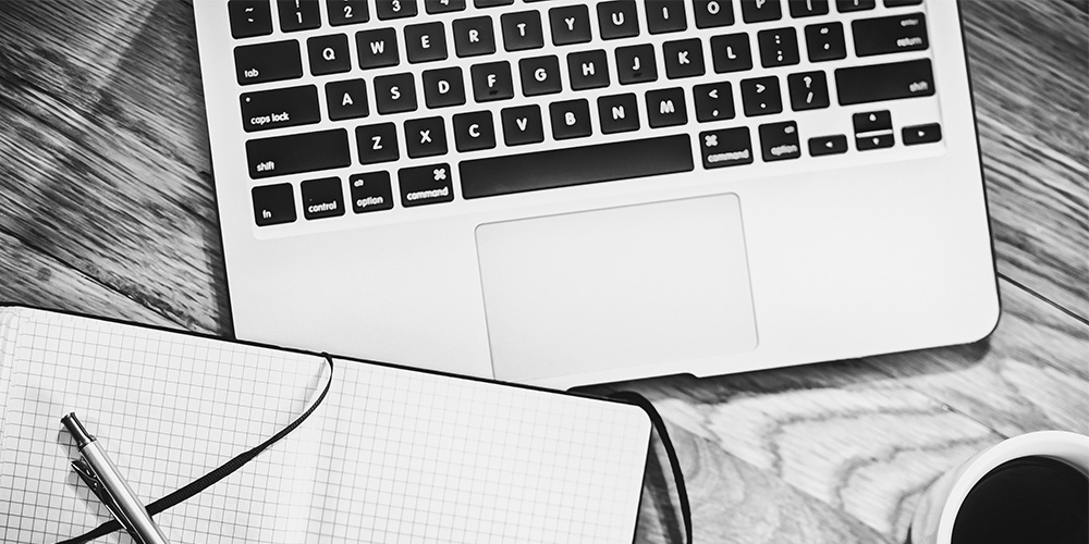 Black and white image showing a laptop keyboard, writing pad, pen and cup of coffee from above