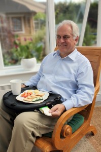 An older man sits in a high-back chair, eating lunch using the Trabasack Curve as a lap tray