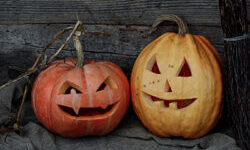 Two pumpkins with scary faces carved into them sat in front of a barn on an old grey blanket with a wooden broom next to them