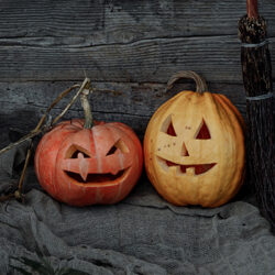 Two pumpkins with scary faces carved into them sat in front of a barn on an old grey blanket with a wooden broom next to them