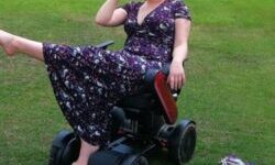 Hannah Barham-Brown in her wheelchair in a park wearing a long black dress with a floral pattern one leg in the air and drinking from a cup