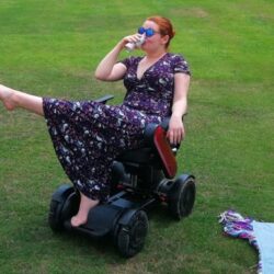 Hannah Barham-Brown in her wheelchair in a park wearing a long black dress with a floral pattern one leg in the air and drinking from a cup