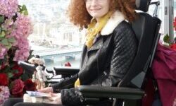 Hannah Deakin with red curly hair in her wheelchair wearing black trousers and a black leather jacket in front of an archway of pink and red flowers