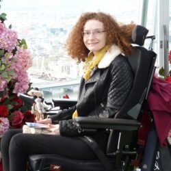 Hannah Deakin with red curly hair in her wheelchair wearing black trousers and a black leather jacket in front of an archway of pink and red flowers