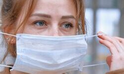 Woman in a beige coat and yellow jumper putting a face mask on