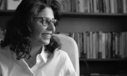 A black and whtie image of Shahd Alshammari with shoulder length hair wearing a white shirt and glasses looking to the side sat in front of a bookcase filled with books