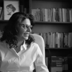 A black and whtie image of Shahd Alshammari with shoulder length hair wearing a white shirt and glasses looking to the side sat in front of a bookcase filled with books