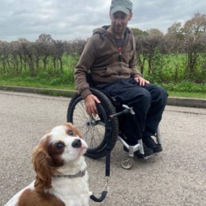 A man sits in a manual wheelchair with a wheelchair dog lead attached to his frame. On the other end of the lead is a very cute dog ready for a walk.